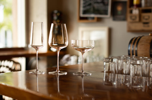 Fable wine glass, larger goblet and coupe glass arranged on a wooden bar counter beside stacked tumblers