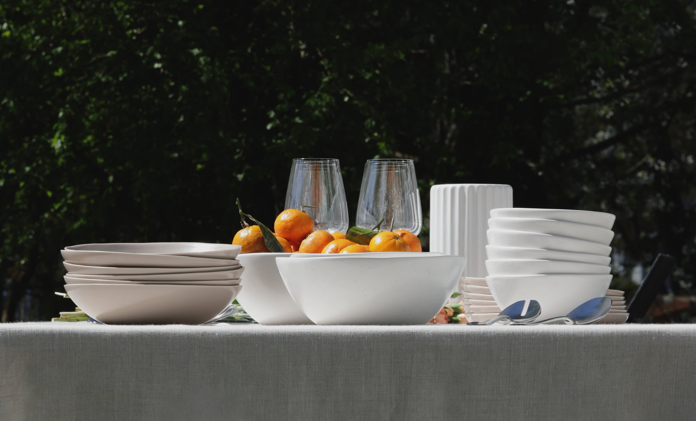 White and stone dinnerware sets, glasses, and cutlery arranged on an outdoor linen-draped table with oranges