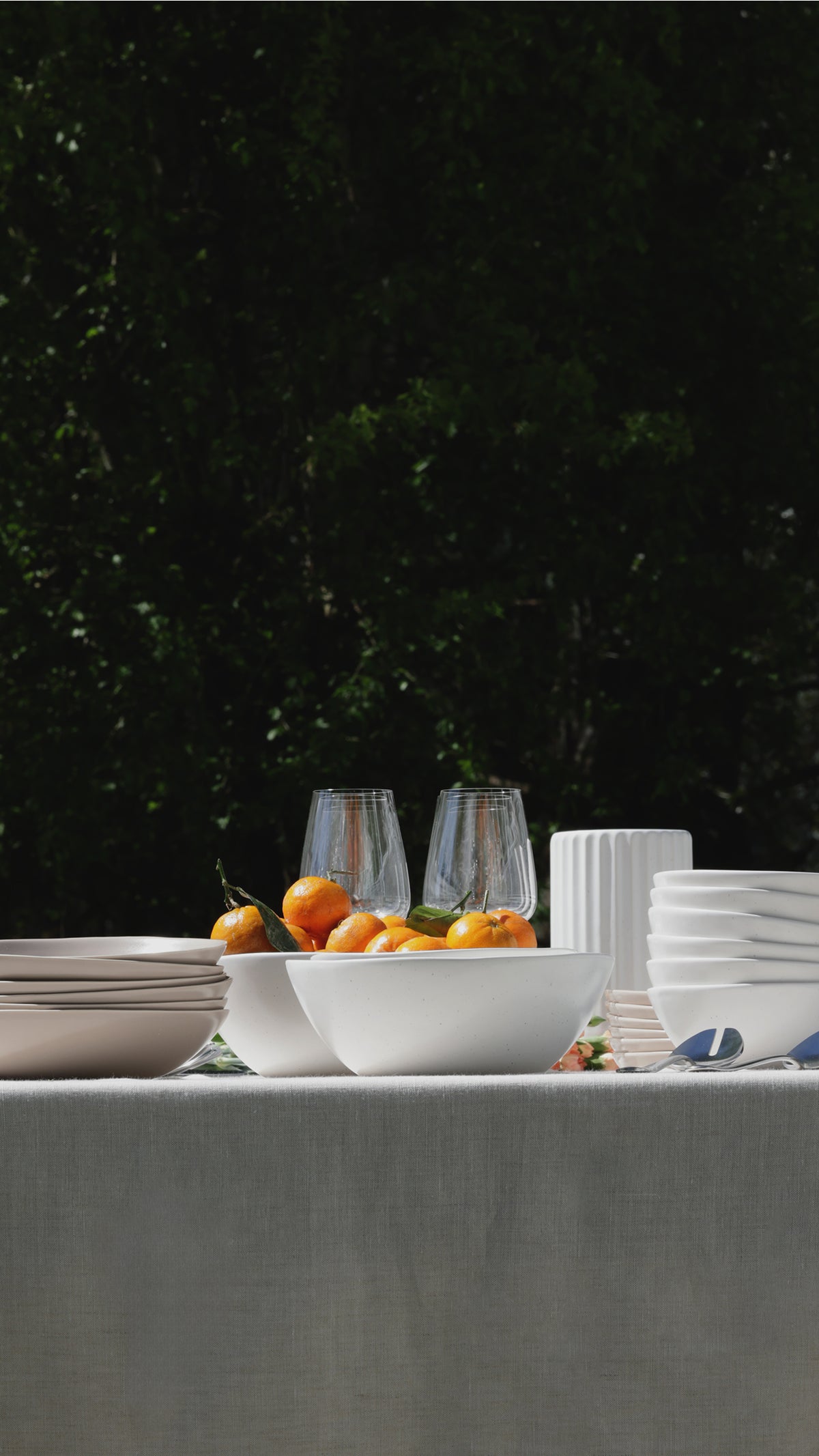 Outdoor table with stacked white dinnerware, a bowl of oranges, and clear glasses against dark green foliage
