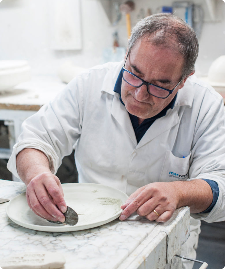 Ceramic artisan in a white coat finishing a Fable plate by hand at a workshop bench
