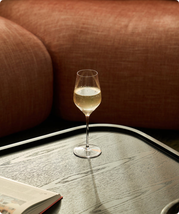 Fable clear wine glass filled with sparkling white wine on a wood tray beside a rust-colored sofa and open book