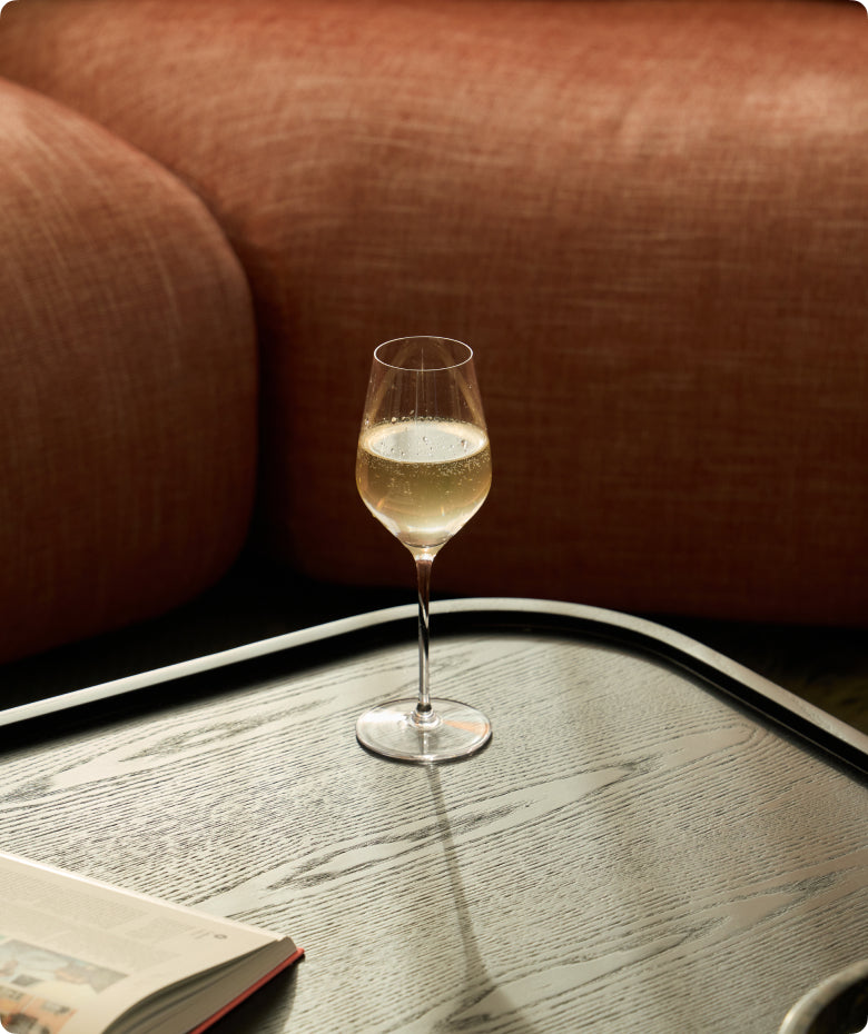 Fable clear wine glass filled with sparkling white wine on a wood tray beside a rust-colored sofa and open book
