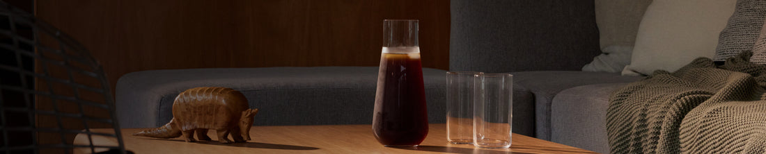 Glass carafe filled with dark coffee drink beside two tall glasses on a wood coffee table in a living room