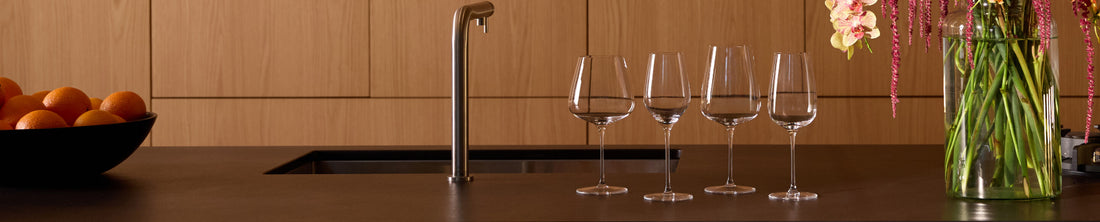 Four clear crystal wine glasses lined up on a dark kitchen countertop beside flowers and oranges