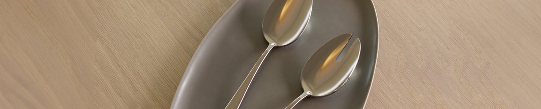 Two stainless steel serving spoons resting on a taupe oval ceramic platter