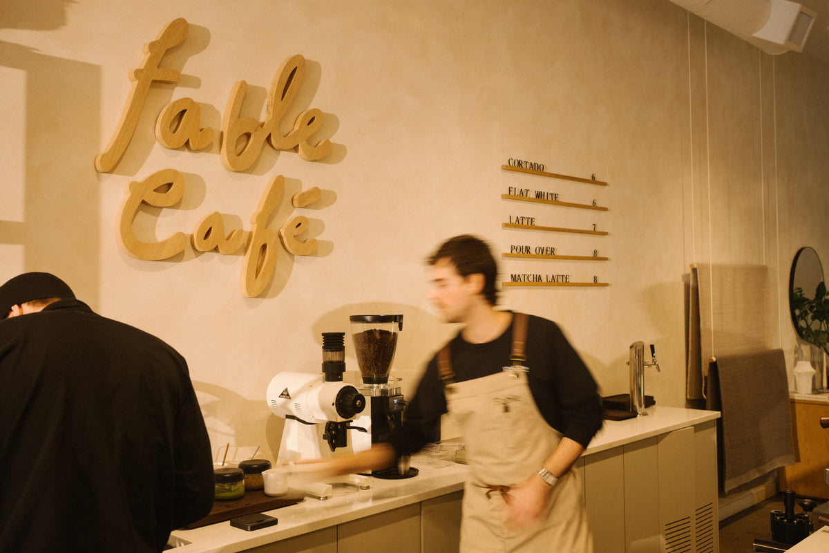 Barista at the Fable Cafe espresso counter beneath a carved Fable Cafe sign and wooden drink menu