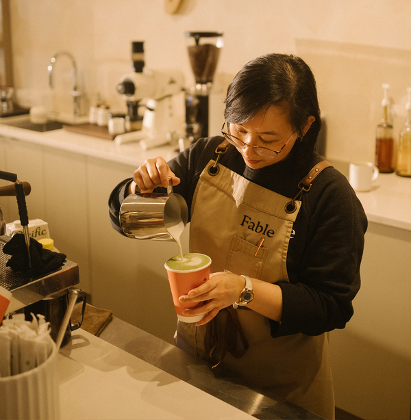 Fable Cafe barista in a branded apron pouring steamed milk into a green matcha latte to-go cup
