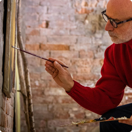 Man in a red sweater painting on an easel against a brick wall.