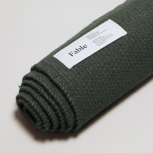 Rolled-up green blanket with a 'Fable' label on the shoreline green wool rug background. #forestgreen